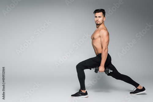 full length of shirtless sportsman working out with dumbbells on grey.