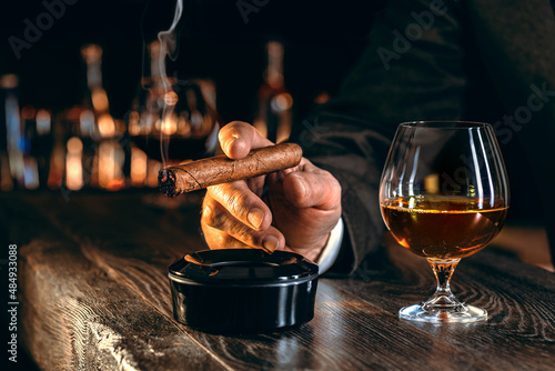 Man\'s hands with a cigar, elegant glass of brandy on the bar counter. Alcoholic drinks, cognac, whiskey, port, brandy, rum, scotch, bourbon. Vintage wooden table in a pub at night