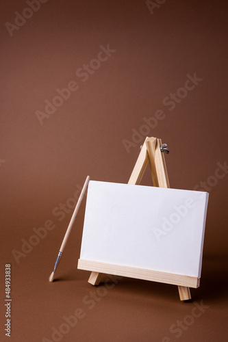 White canvas on a small wooden easel stand minimal concept on the brown backdrop. Empty blank mock up frame for design with copy space. 