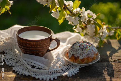 Pottery cup with milk (kefir, yogurt, sour cream, kumis), cupcake, lace tablecloth, wooden table. Outdoor picnic, breakfast, brunch, refreshments. Soft focus