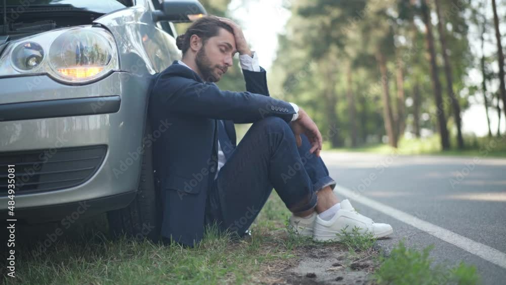 Side view hopeless young man sitting at car breakdown lights on with vehicle passing on suburban road. Sad stressed Caucasian male driver outdoors on sunny day thinking. Accident concept Side view hopeless young man sitting at car breakdown lights on with vehicle passing on suburban road. Sad stressed Caucasian male driver outdoors on sunny day thinking. Accident concept
