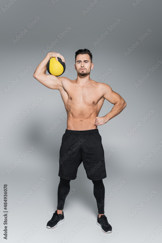 full length of shirtless and bearded sportsman holding ball and standing with hand on hip on grey.