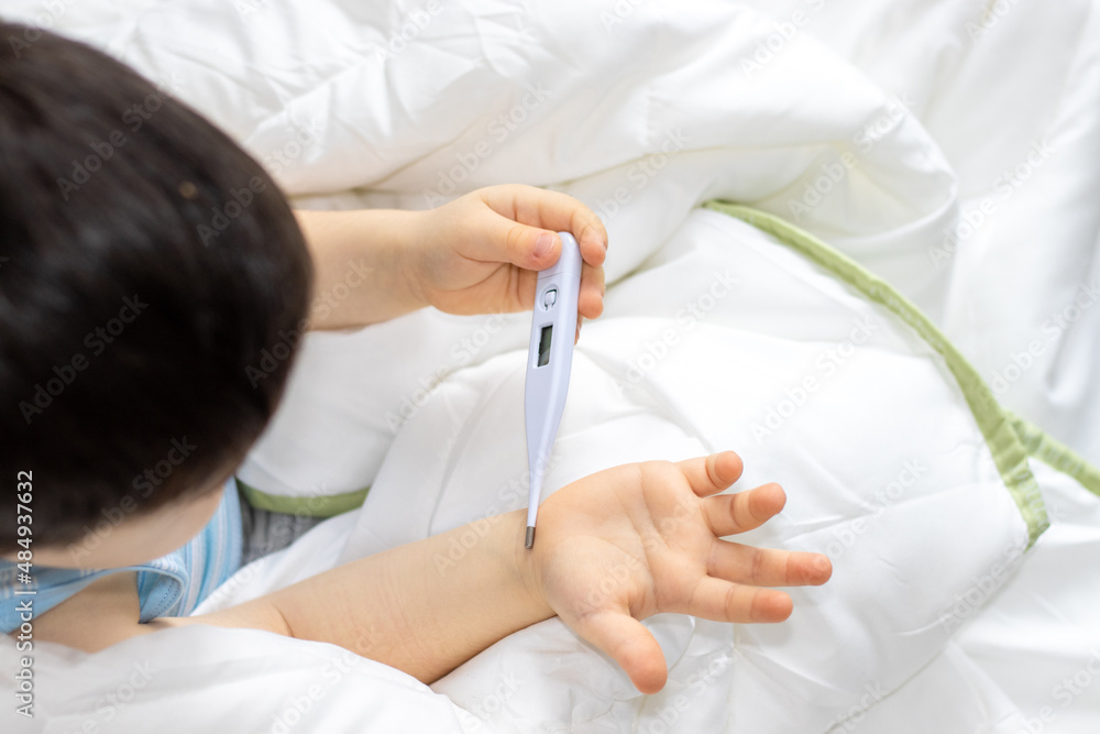 cute little kid in t shirt laying on the bed or is sitting with the thermometer in hand and looking at it. boy wrapped in white soft blanket measure feaver, on the bed. cold, fever. virus,kindergarden cute little kid in t shirt laying on the bed or is sitting with the thermometer in hand and looking at it. boy wrapped in white soft blanket measure feaver, on the bed. cold, fever. virus,kindergarden