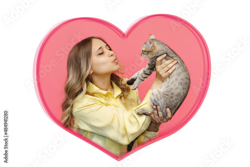 A young caucasian cute blonde woman in a yellow shirt holds a cat in her hands admiring it inside a heart-shaped frame on a pink background. The girl babying with a kitten. Fiendship of pet and owner