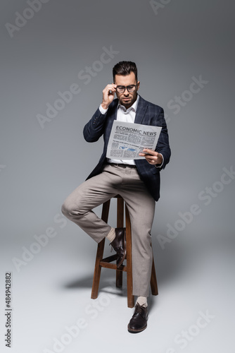 full length of bearded businessman in blazer and glasses reading newspaper and sitting on chair on grey. © LIGHTFIELD STUDIOS