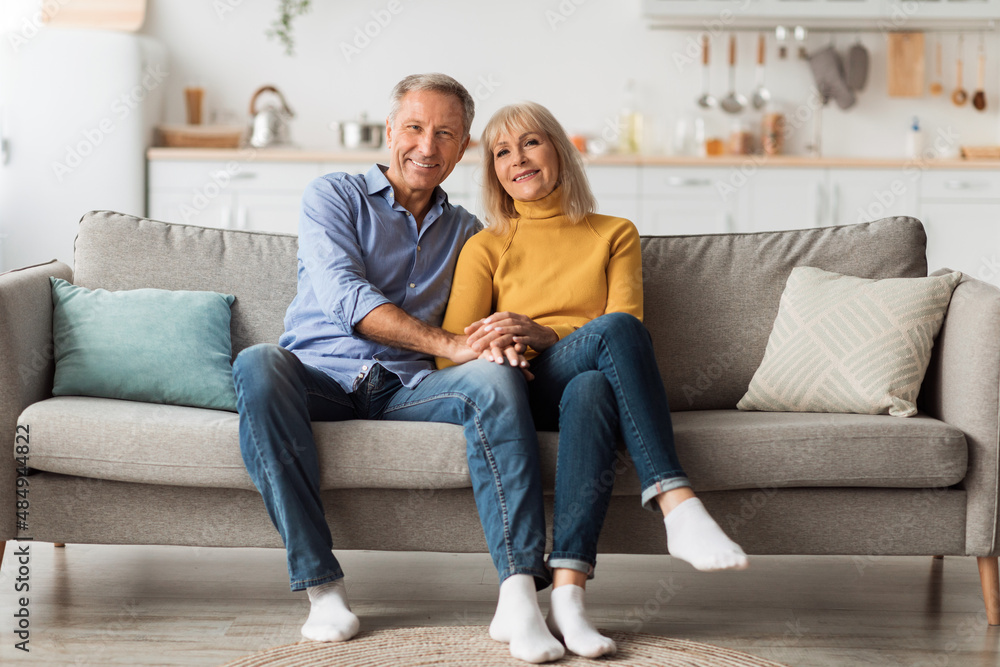 Cheerful Senior Spouses Holding Hands Hugging Sitting On Sofa Indoor