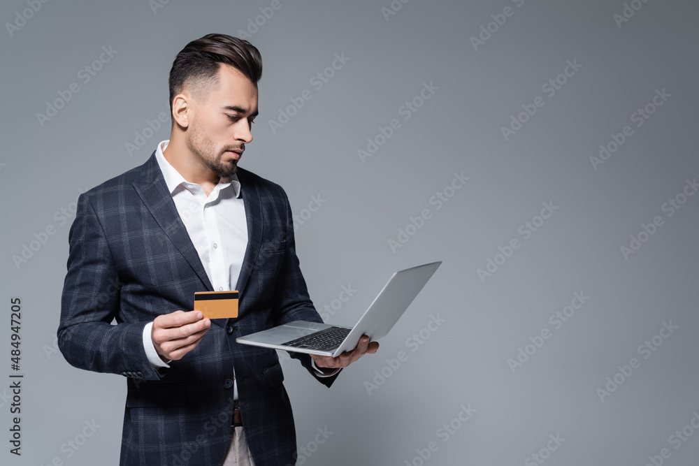 businessman in suit holding credit card and using laptop isolated on grey. businessman in suit holding credit card and using laptop isolated on grey.