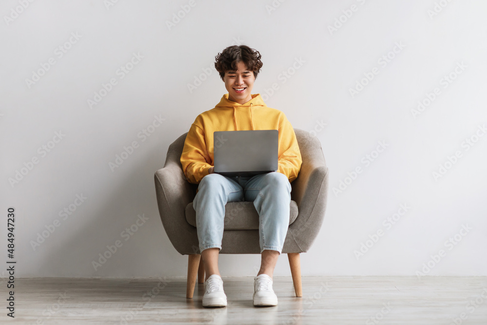 Happy young Asian male working online, sitting in armchair and using laptop against white studio wall, full length