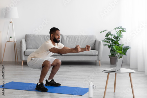 Concentrated young black bearded athlete man in white sports clothes squatting in living room interior, profile
