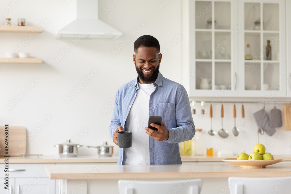 Cheerful young black male with cup of coffee in hand in minimalist kitchen interior reads news on smartphone Cheerful young black male with cup of coffee in hand in minimalist kitchen interior reads news on smartphone