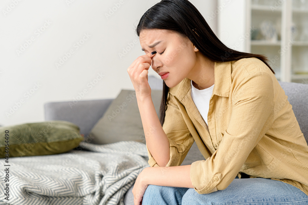 Sad asian woman feels bad, sits on the sofa at home, holding head and closed her eyes. Upset asian lady has problem and depression, has poor health, anxiety Sad asian woman feels bad, sits on the sofa at home, holding head and closed her eyes. Upset asian lady has problem and depression, has poor health, anxiety