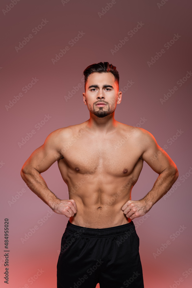 lighting on strong man with muscles posing with hands on hips on pink and grey.