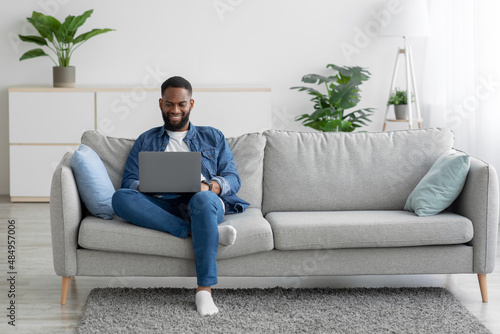 Quadro su tela Smiling millennial black bearded guy working on laptop on sofa in living room in