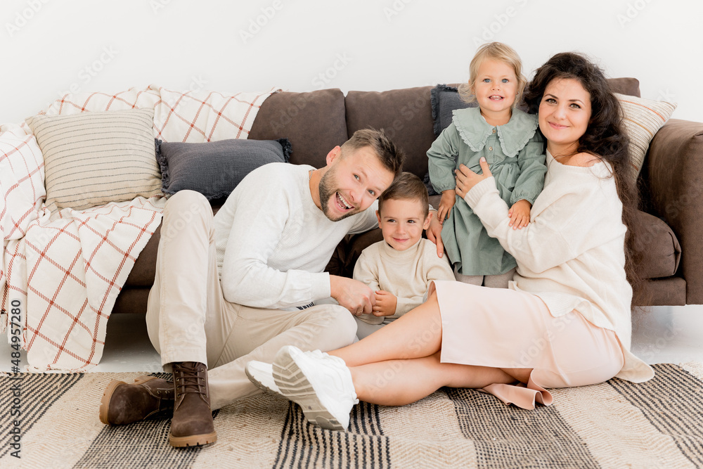 Happy beautiful parents sitting near the sofa with joyful little kids, smiling, spending weekends at home. Charming father with pretty mother playing with cute daughter and son, family concept Happy beautiful parents sitting near the sofa with joyful little kids, smiling, spending weekends at home. Charming father with pretty mother playing with cute daughter and son, family concept