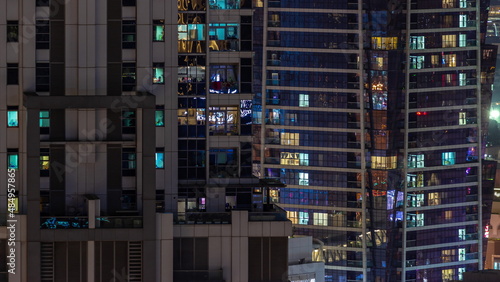 Apartment windows of a glazed skyscraper glow at night with city lights reflection aerial timelapse.