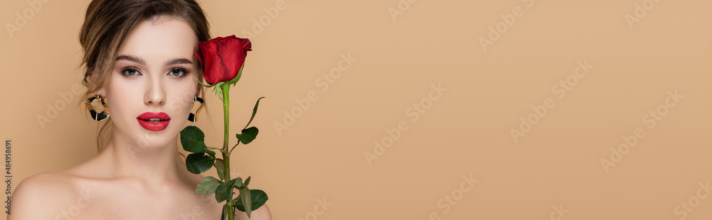 pretty woman with makeup and bare shoulders looking at camera near red rose isolated on beige, banner.