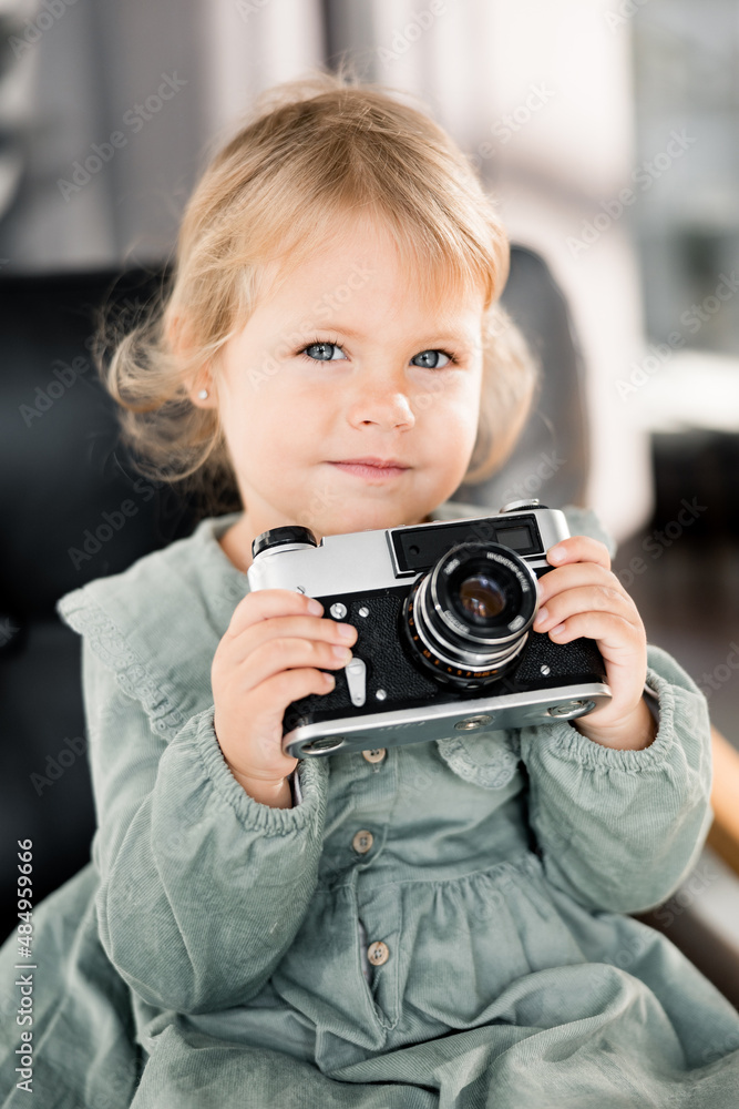 Portrait of adorable cute baby girl holding in arms little photo camera, smiling, wearing stylish green dress. Beautiful lovely toddler sitting on chair and looking at the camera