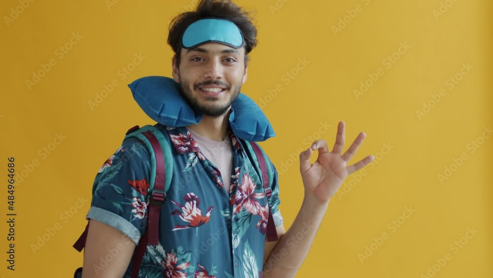 Middle Eastern man tourist with backpack travel pillow and sleep mask showing ok hand gesture and inviting to join on trip during vacation Middle Eastern man tourist with backpack travel pillow and sleep mask showing ok hand gesture and inviting to join on trip during vacation