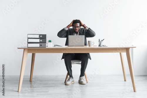 Stressed young black man using laptop, sitting at desk, grabbing his head, having problem performing work task