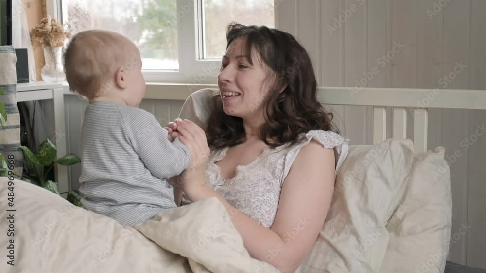 One Caucasian brunette mother in nightwear waking up with her tiny newborn, playing on bed. Young woman with baby relaxing at home. Bed-sharing concept. Infant child with his mom in bedroom. One Caucasian brunette mother in nightwear waking up with her tiny newborn, playing on bed. Young woman with baby relaxing at home. Bed-sharing concept. Infant child with his mom in bedroom.