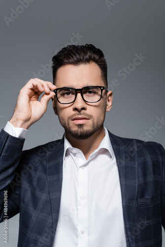 bearded businessman in blazer adjusting glasses isolated on grey.