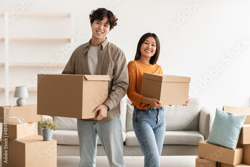 Portrait of young Asian woman and her cool boyfriend holding cardboard boxes, looking at camera and smiling in new home © Prostock-studio