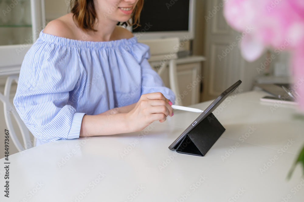 Fototapeta premium Young brunette woman designer in stylish blouse develops new clothes via tablet on stand at white table near flowers at home close view