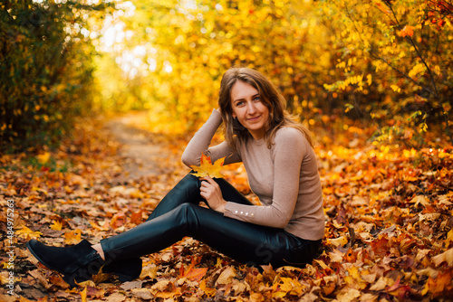 Beautiful girl is sitting on yellow leaves in the park. Attractive woman wearing beige sweater and black pants is walking in autumn park. Bright, warm sunny autumn day with yellow background. 