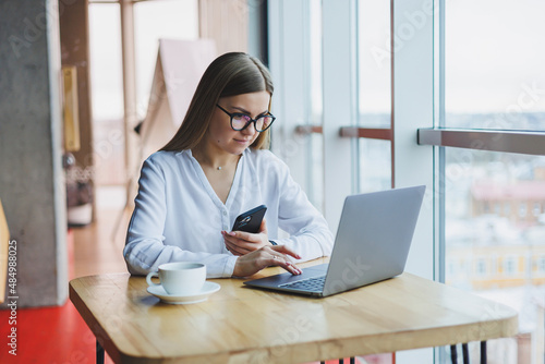 A smart woman manager with a cup of coffee has a business conversation via a smartphone to discuss a delivery order, a young woman small owner sits at a table in a cafe with a laptop and calls