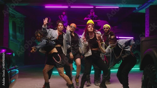 Group of stylish young people dancing , moving near military tank inside garage building Colorful stylish clothes , hippie style fashion . Modern dances . Purple led light , slow motion