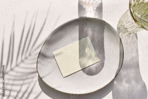 Eco minimal tableware scene.  Empty blank on ceramic plate, cocktail glass on neutral background. Sunlight, palm shadow.  Mockup invitation card for wedding, business or menu. Wabi sabi style