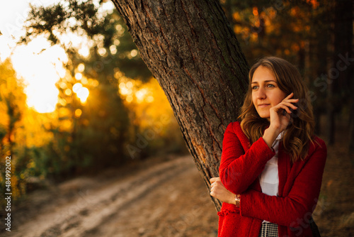Elegant young girl at sunset in the forest. Stylish girl in a red coat and skirt. Warm autumn in the forest. Magic sunset and portrait of a girl  © Anhelina Tyshkovets