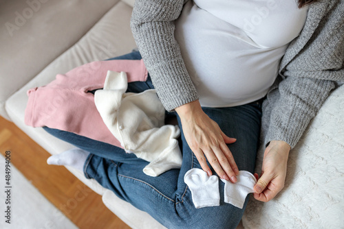 Top-down view of an unrecognizable Caucasian woman in third trimester of pregnancy sorting through baby clothing on a sofa