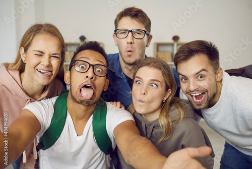 Happy students taking funny selfie indoors.Group of joyful young multiethnic friends having fun together, taking selfie on mobile phone, making hilarious grimaces, sticking tongues out, squinting eyes photo