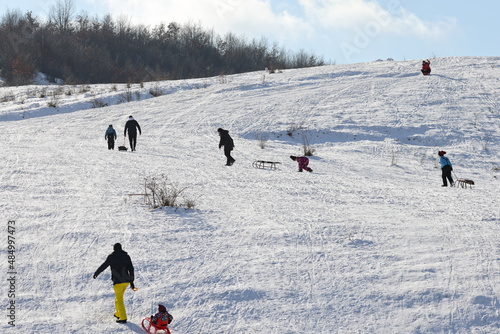 Recreation people and children go sledding, skiing on the sunny, snowy hillside on the weekends in winter. Color landscape photo. Illustration of winter sports, holidays and tourism.