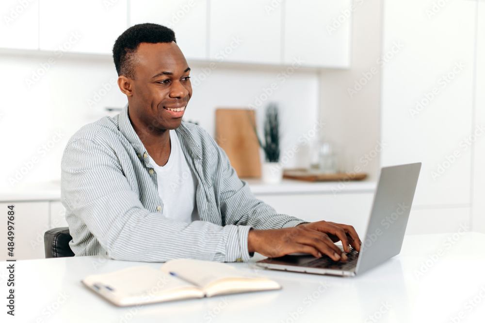 Attractive positive african american guy, freelancer, designer, IT specialist, works on a laptop at home, develops a strategy, analyzes, messaging with clients or colleagues, browsing internet, smiles Attractive positive african american guy, freelancer, designer, IT specialist, works on a laptop at home, develops a strategy, analyzes, messaging with clients or colleagues, browsing internet, smiles