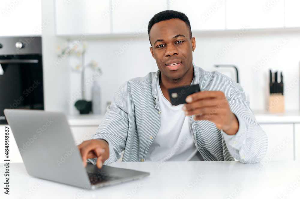 Positive African American young adult male, sitting at a table in the kitchen, using a computer and a bank card to pay for purchases and deliveries online, entering data, looking at a card, smiling. Positive African American young adult male, sitting at a table in the kitchen, using a computer and a bank card to pay for purchases and deliveries online, entering data, looking at a card, smiling.