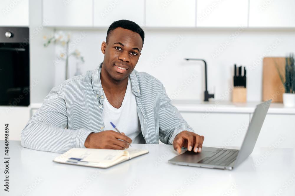 Attractive African American guy, student, freelancer, sits at a table in the kitchen, uses laptop and notepad for online learning, looks at the camera, listens to a lecture, takes notes,smile friendly Attractive African American guy, student, freelancer, sits at a table in the kitchen, uses laptop and notepad for online learning, looks at the camera, listens to a lecture, takes notes,smile friendly