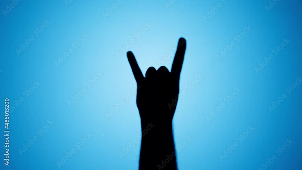 Man showing rock and roll gesture with fingers isolated on blue background. Male person making shadow silhouette with hand close-up. Man showing rock and roll gesture with fingers isolated on blue background. Male person making shadow silhouette with hand close-up.