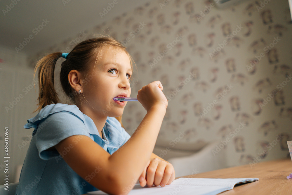 Close-up of thinking primary little child girl doing homework and holding pen against mouth sitting at home table by window. Pensive dreaming preschool kid studying at bedroom. Homeschooling concept. Close-up of thinking primary little child girl doing homework and holding pen against mouth sitting at home table by window. Pensive dreaming preschool kid studying at bedroom. Homeschooling concept.
