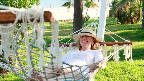 senior woman in straw hat smiling happy relaxing on a hammock enjoying the fresh air on the terrace around palm trees near the sea. Senior citizen lifestyle concept