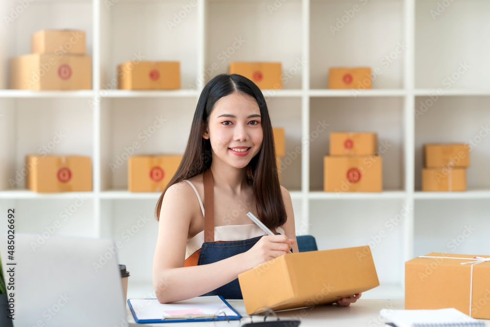 Portrait of young Asian small business owner working at home office writing address on cardboard box at workplace. Smile woman is working at warehouse for online store looking at camera. Portrait of young Asian small business owner working at home office writing address on cardboard box at workplace. Smile woman is working at warehouse for online store looking at camera.
