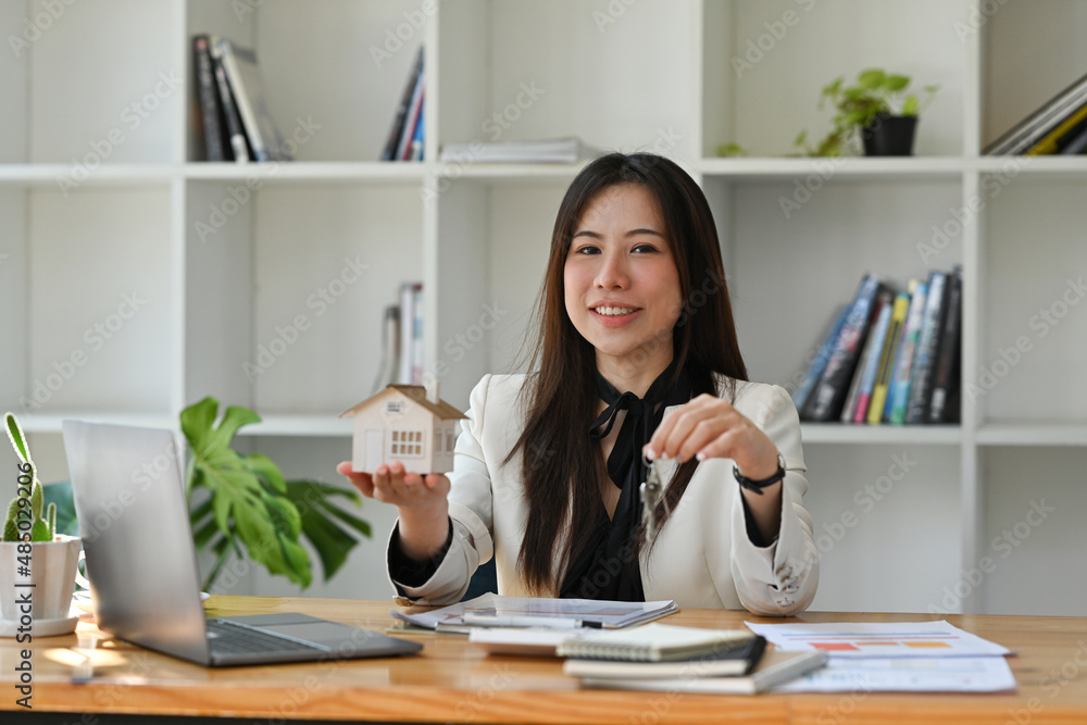 Smiling asian woman holding house model and house key in hand. Real estate investment and insurance concept.