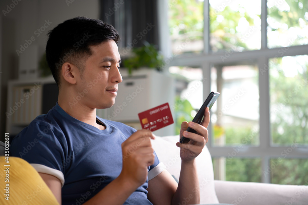 Asian man sitting comfortably at home enjoying and browsing online shopping and easy and convenient payment using credit card with online payment and banking application on smartphone. Asian man sitting comfortably at home enjoying and browsing online shopping and easy and convenient payment using credit card with online payment and banking application on smartphone.