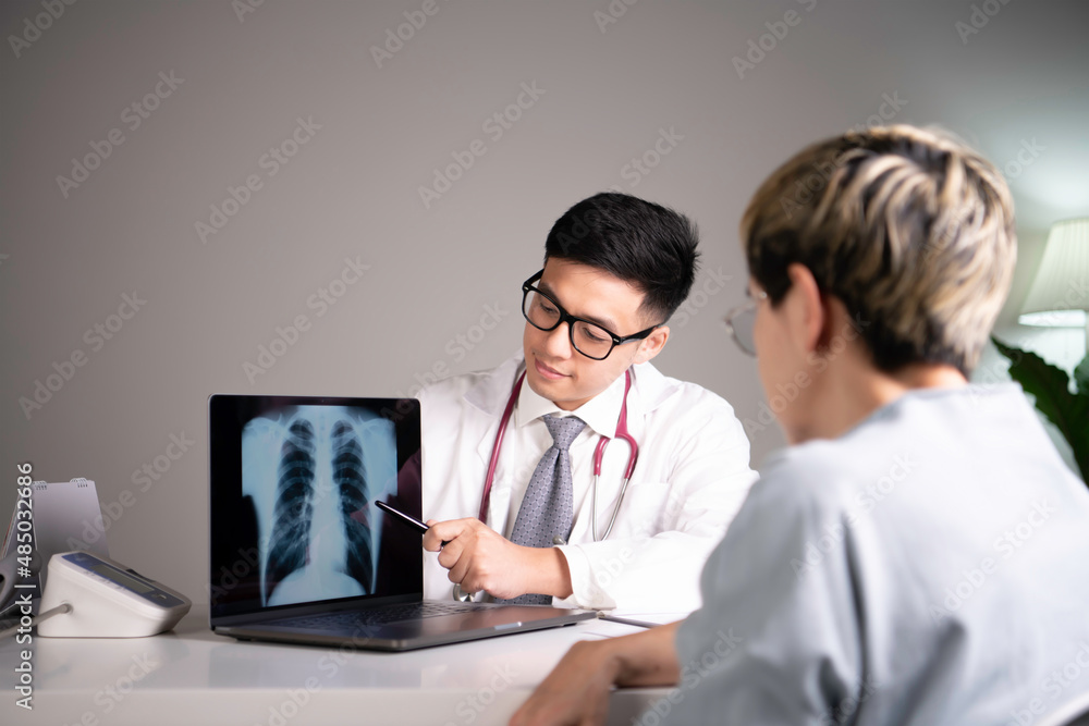 Asian medical doctor orthopaedic surgeon wearing stethoscope showing x-ray image from laptop while explaining and soothing patient about diagnosis and treatment plan in examination room in hospital. Asian medical doctor orthopaedic surgeon wearing stethoscope showing x-ray image from laptop while explaining and soothing patient about diagnosis and treatment plan in examination room in hospital.