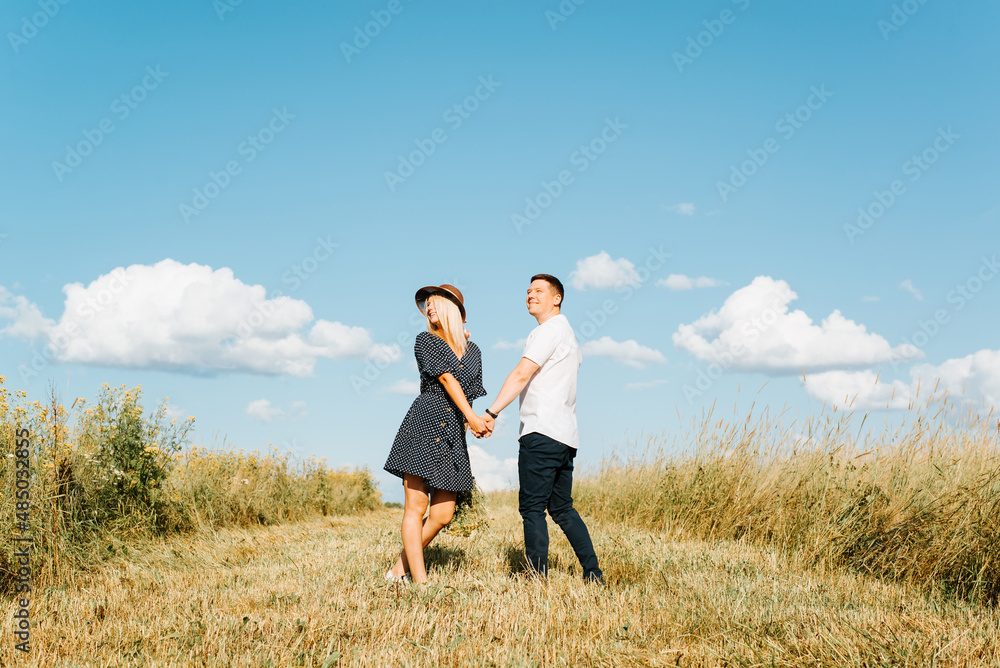 Happy loving couple holding hands together while walking on field and looking away, outdoors. Cute young caucasian family spending time with each other in nature on sunny summer day