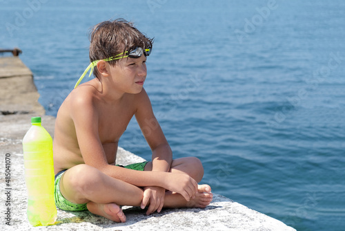 A cute boy in swimming glasses and shorts with a bottle of water sits on a pier on the seashore. photo