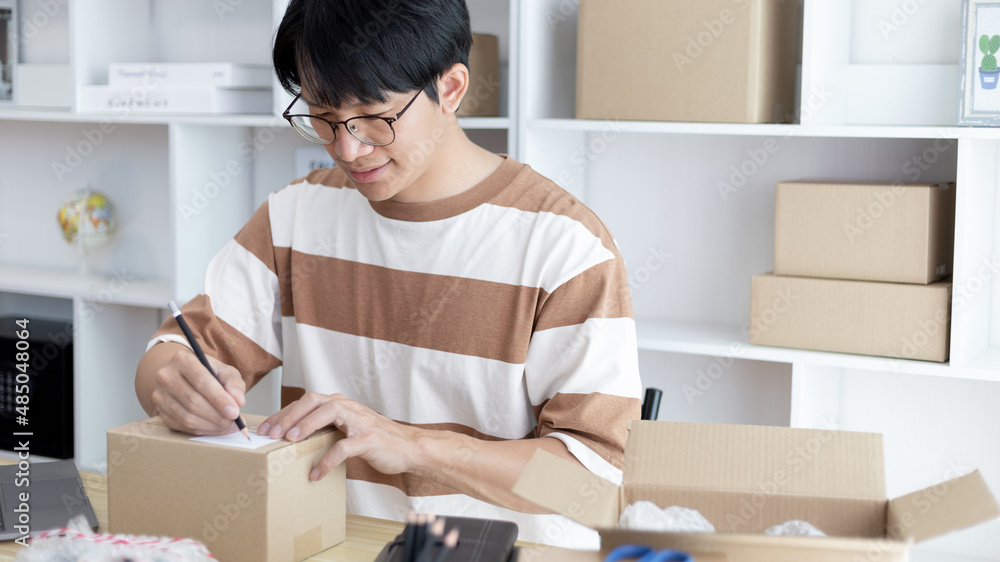 Young Asian man is writing down the customer's details and addresses on the notebook or box in order to prepare for shipping according to the information, packing box, Sell online, freelance working. Young Asian man is writing down the customer's details and addresses on the notebook or box in order to prepare for shipping according to the information, packing box, Sell online, freelance working.