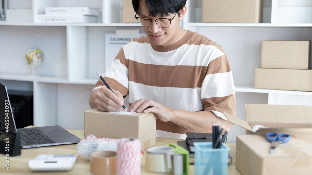 Young Asian man is writing down the customer's details and addresses on the notebook or box in order to prepare for shipping according to the information, packing box, Sell online, freelance working. Young Asian man is writing down the customer's details and addresses on the notebook or box in order to prepare for shipping according to the information, packing box, Sell online, freelance working.