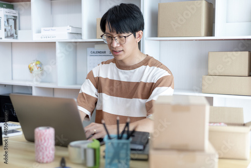 Young man uses a laptop to chat with customers who come to order product, Freelance work at home, Packaging on background, Sell online, Small business owner, Online shopping SME entrepreneur.